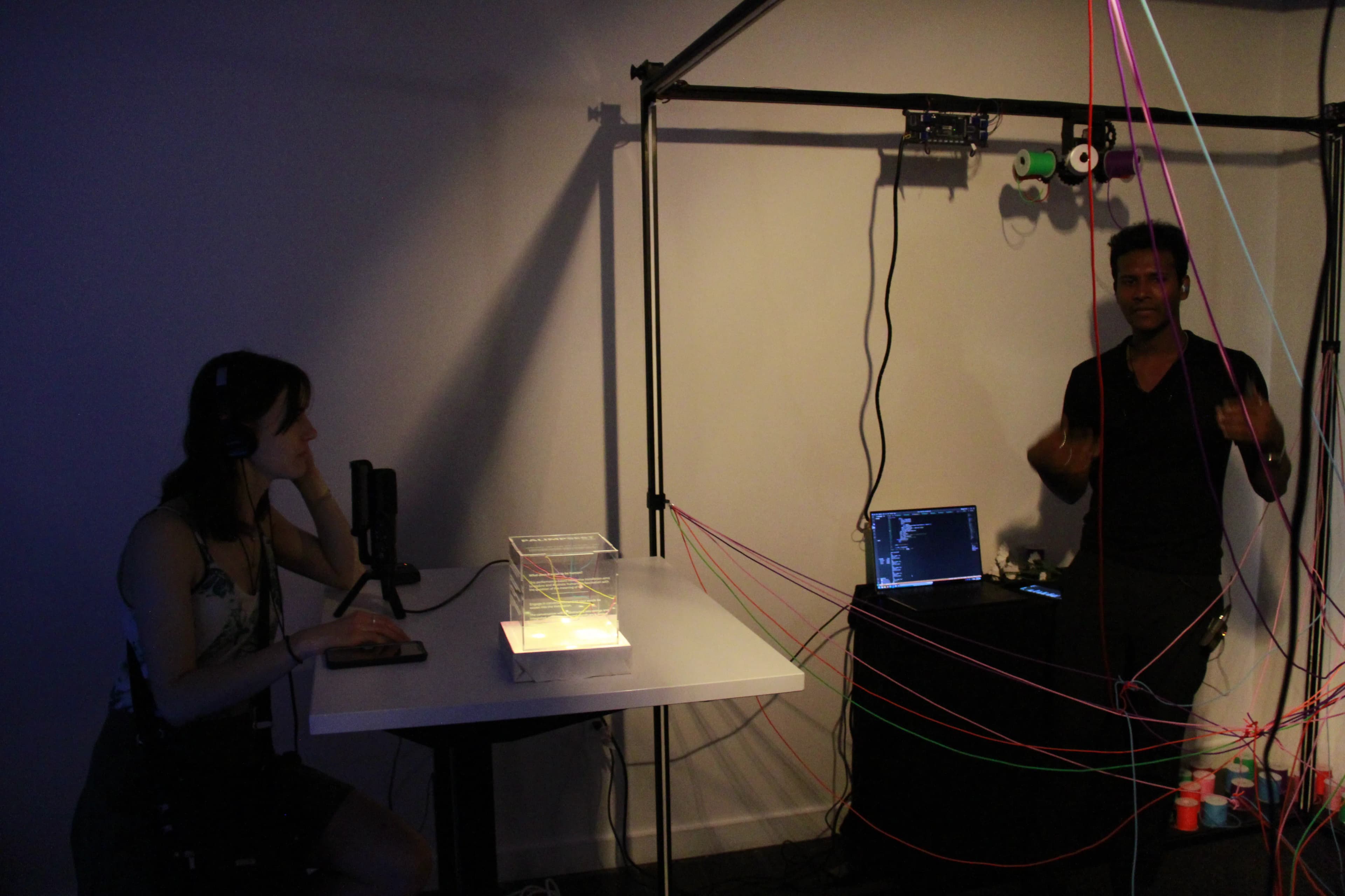 A participant converses into a microphone while Leffin places threads inside the metal cube frame